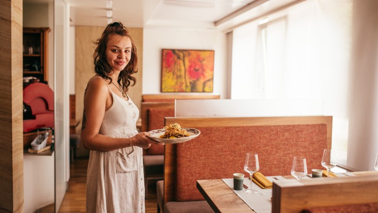 A woman in a restaurant holds a plate of food. She is wearing a white dress and smiling at the camera.