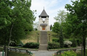 A stone war memorial with a tower and steps, surrounded by trees and benches.