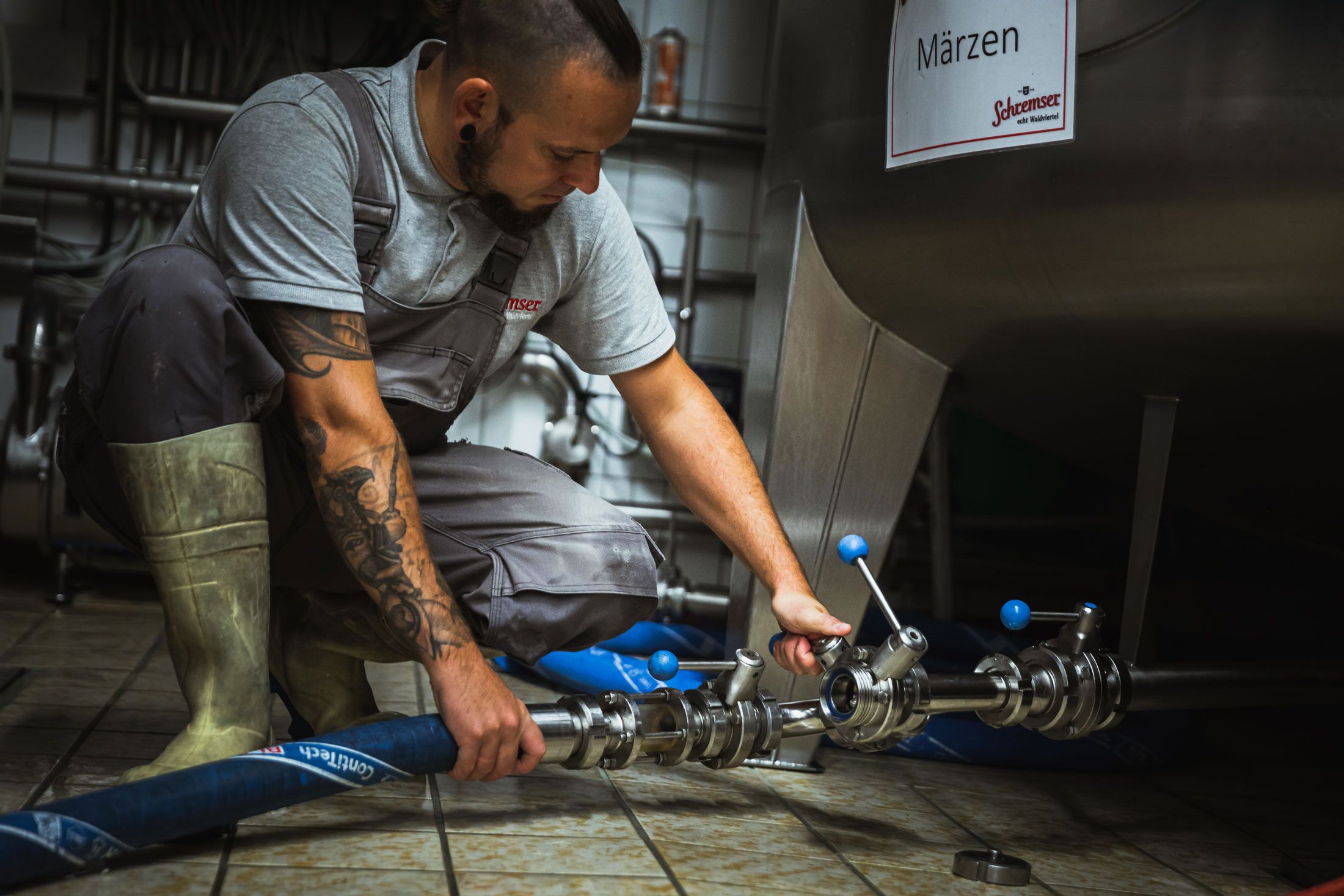 A man in work clothes holds glasses of beer in front of a brewery.