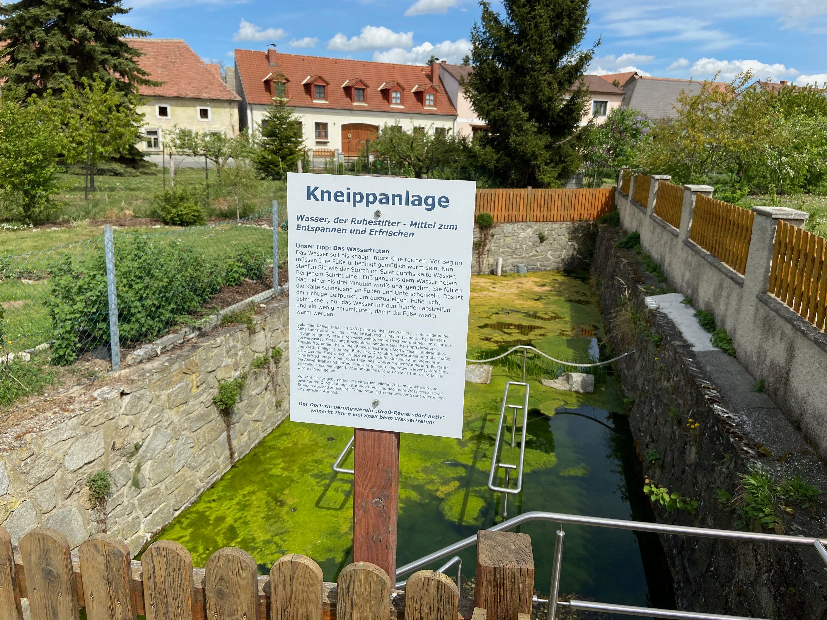 Kneipp facility with information board and water basin, surrounded by a fence and houses in the background.