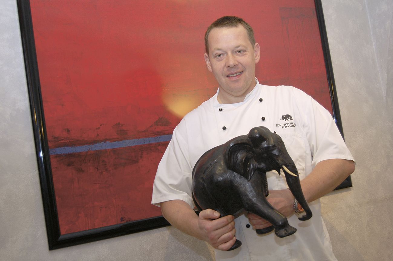A cook in a white uniform holds a black elephant figure in front of a red painting.
