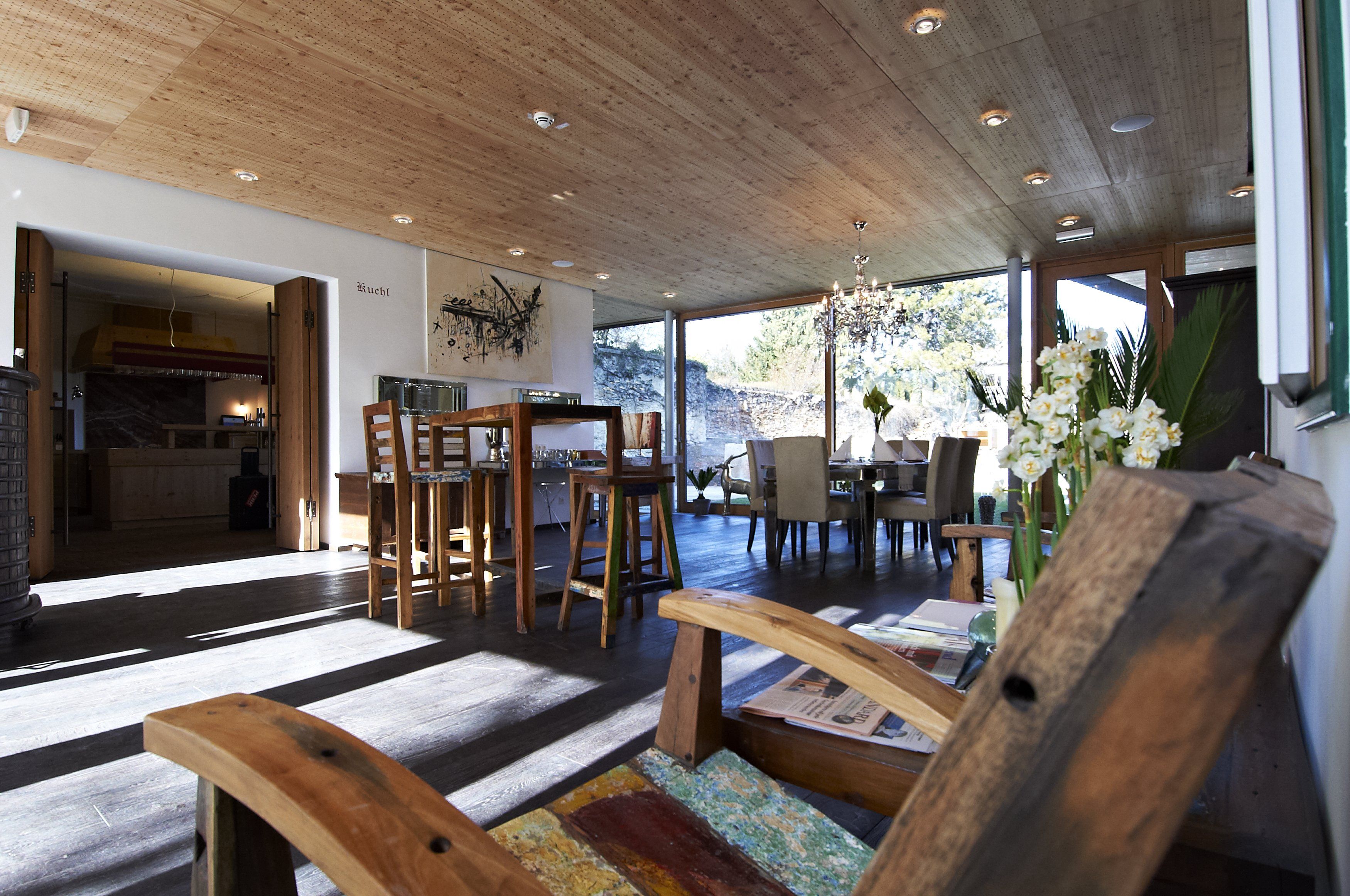 A modern breakfast room with wooden furniture, large windows and a chandelier. A wooden chair in the foreground and a dining table in the background.