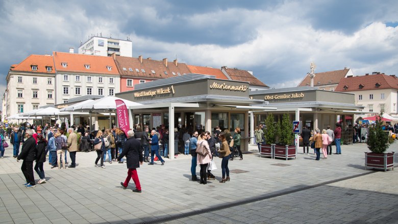 Crowd on a busy marketplace with stalls and buildings in the background.