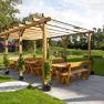 Wooden pergola with benches and tables outside, surrounded by lawn and trees.