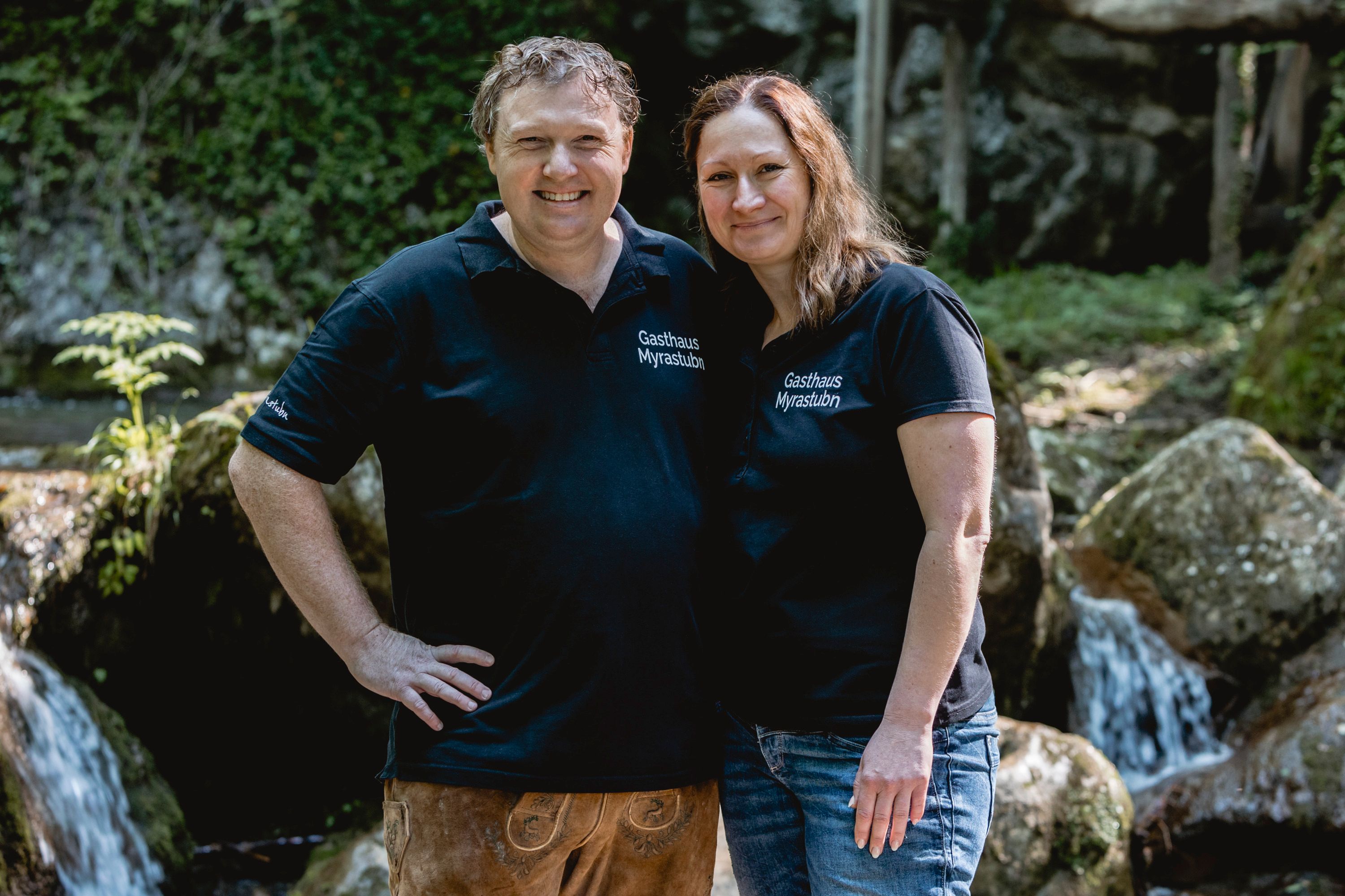 A man and a woman in black T-shirts with the inscription 'Gasthaus Myrastubn' stand smiling in front of a waterfall.