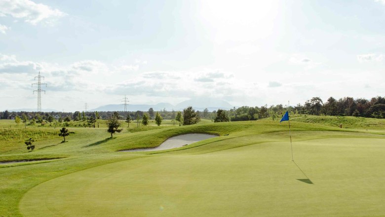 Golf course with flag on the green, surrounded by trees and electricity pylons under a cloudy sky.