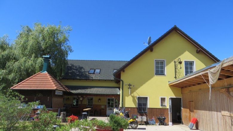 Gasthof Oase am Teich, © Urani Yellow building with green roof and terrace, surrounded by plants, under a blue sky.