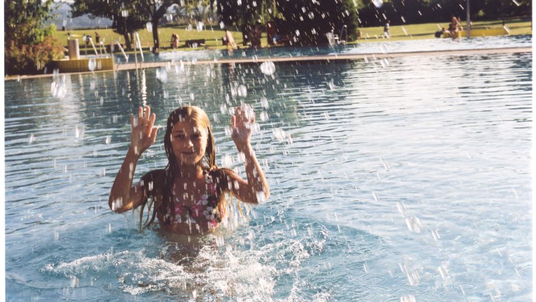 A girl in the water of an outdoor pool, surrounded by splashing water, with trees and meadow in the background.