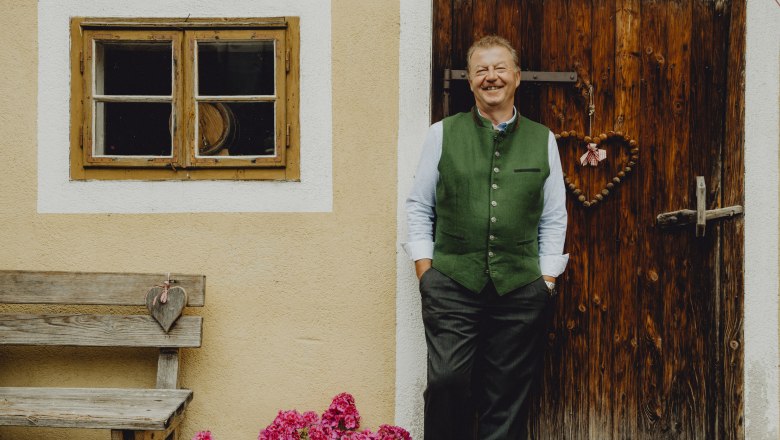 A man in traditional dress stands smiling in front of a wooden door next to a window and a bench with flowers.