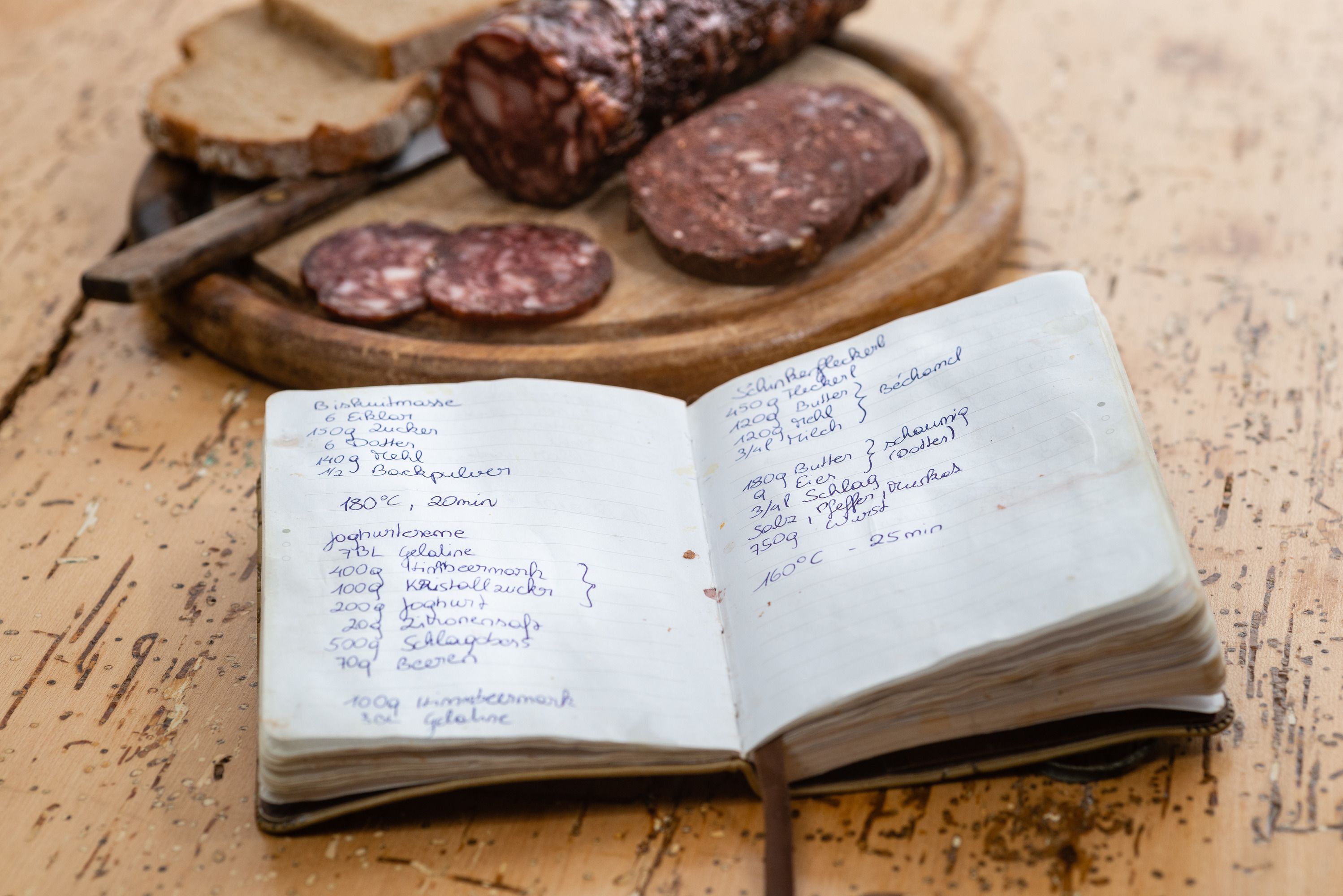 A handwritten recipe book lies on a table next to a wooden platter with venison sausage and bread.