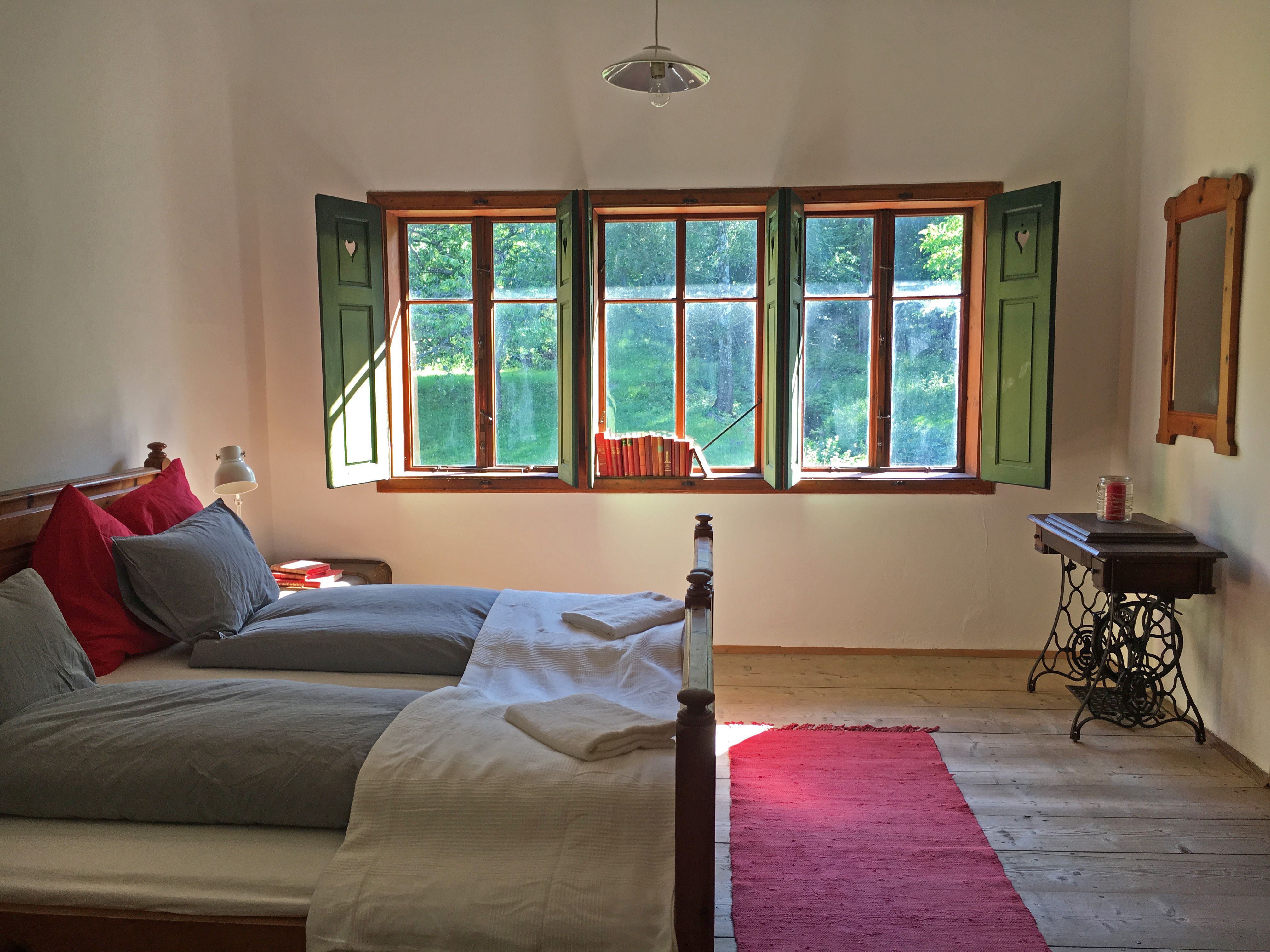 Cozy double room with wooden floor, bed, window with green shutters and a red carpet.