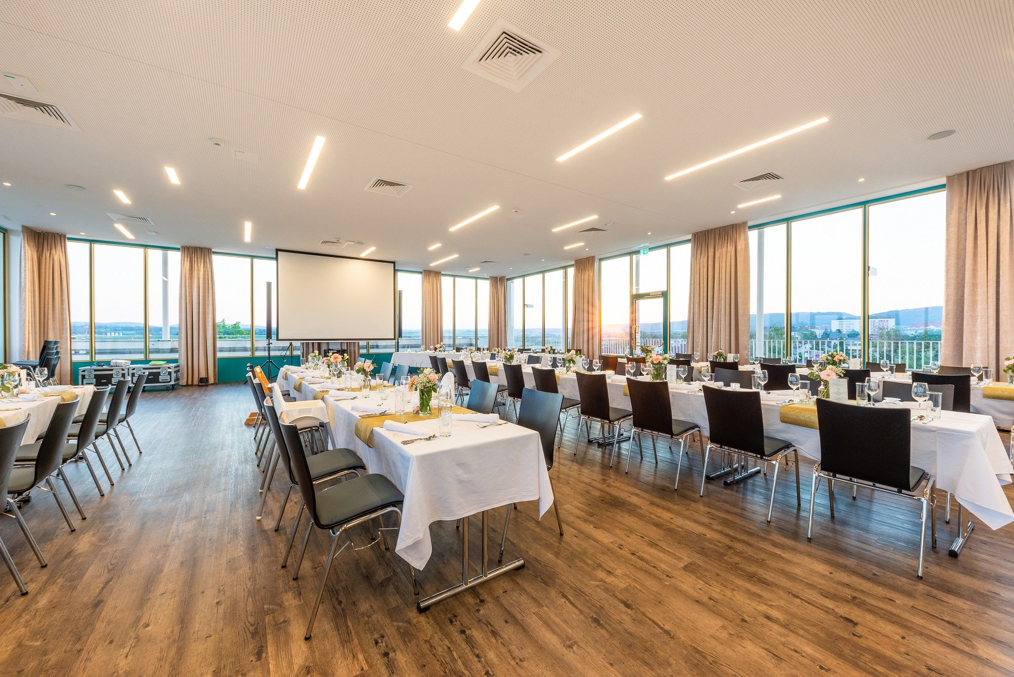 A large, bright room with covered tables and chairs, large windows and a projector.