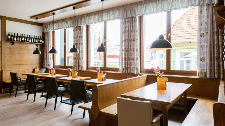 Cozy guest room with wooden tables, chairs and benches, decorated with flowers and menus. Large windows with curtains and hanging lamps.