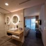 Modern hotel room with open bathroom, washbasin, mirror and shower. In the background a bed and a window with curtains.