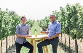 Two men sit at a table in the vineyard with wine glasses.
