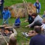 People sit around a campfire and roast bread on long sticks.