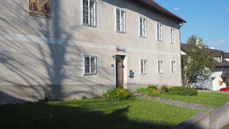 A two-story building with several windows and a sign above the door, surrounded by a well-tended garden.