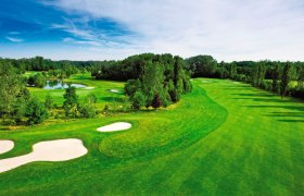 Green golf course with bunkers and pond, surrounded by trees under a blue sky.