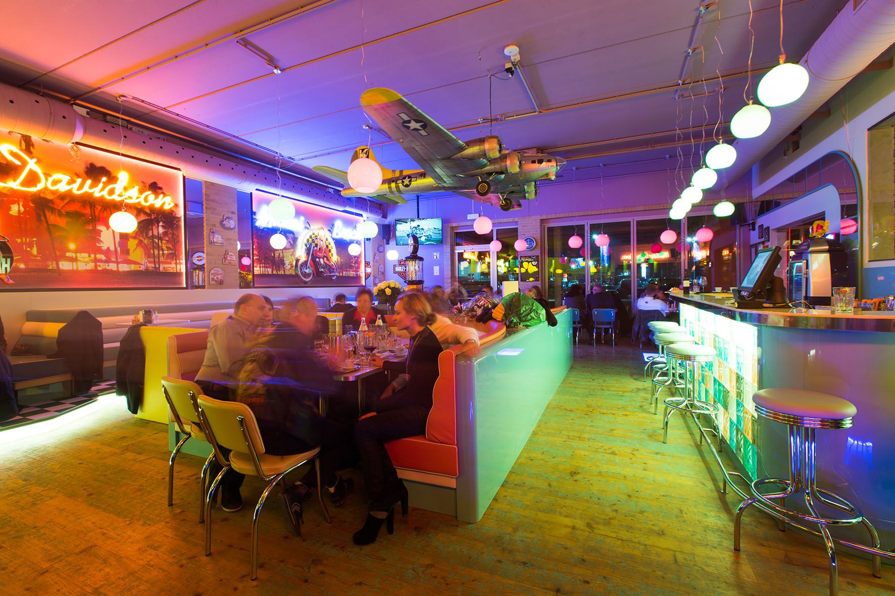 Interior view of a colorfully lit diner with bar, tables and an airplane model on the ceiling.