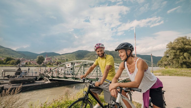 Two cyclists with helmets stand smiling in front of a ferry in a rural landscape.