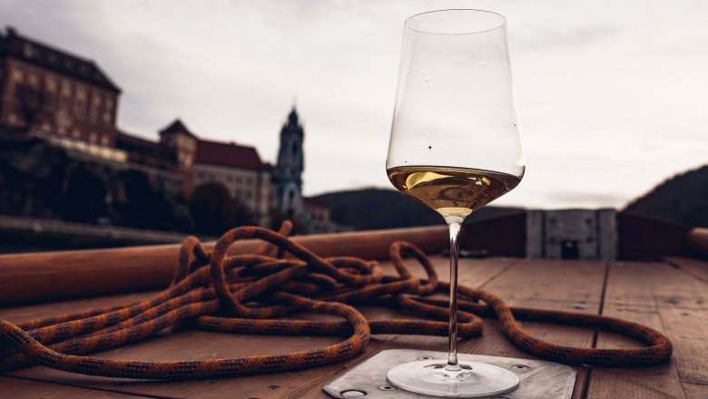 A wine glass with white wine stands on a boat, the church of D&uuml;rnstein is blurred in the background.