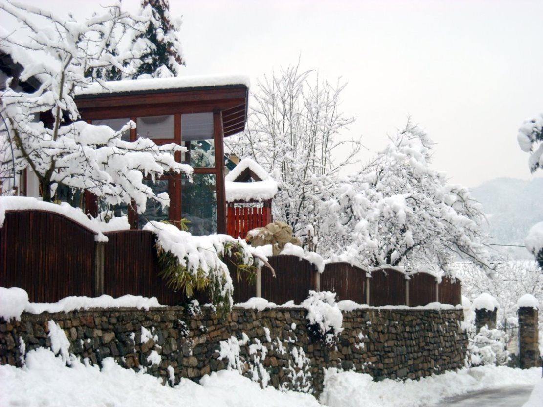 A snow-covered house with a wooden fence and stone wall, surrounded by snow-covered trees.