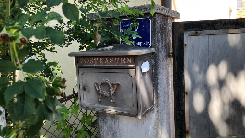 An old letterbox with the inscription 'Postbox' on a wall next to a gate, surrounded by plants.
