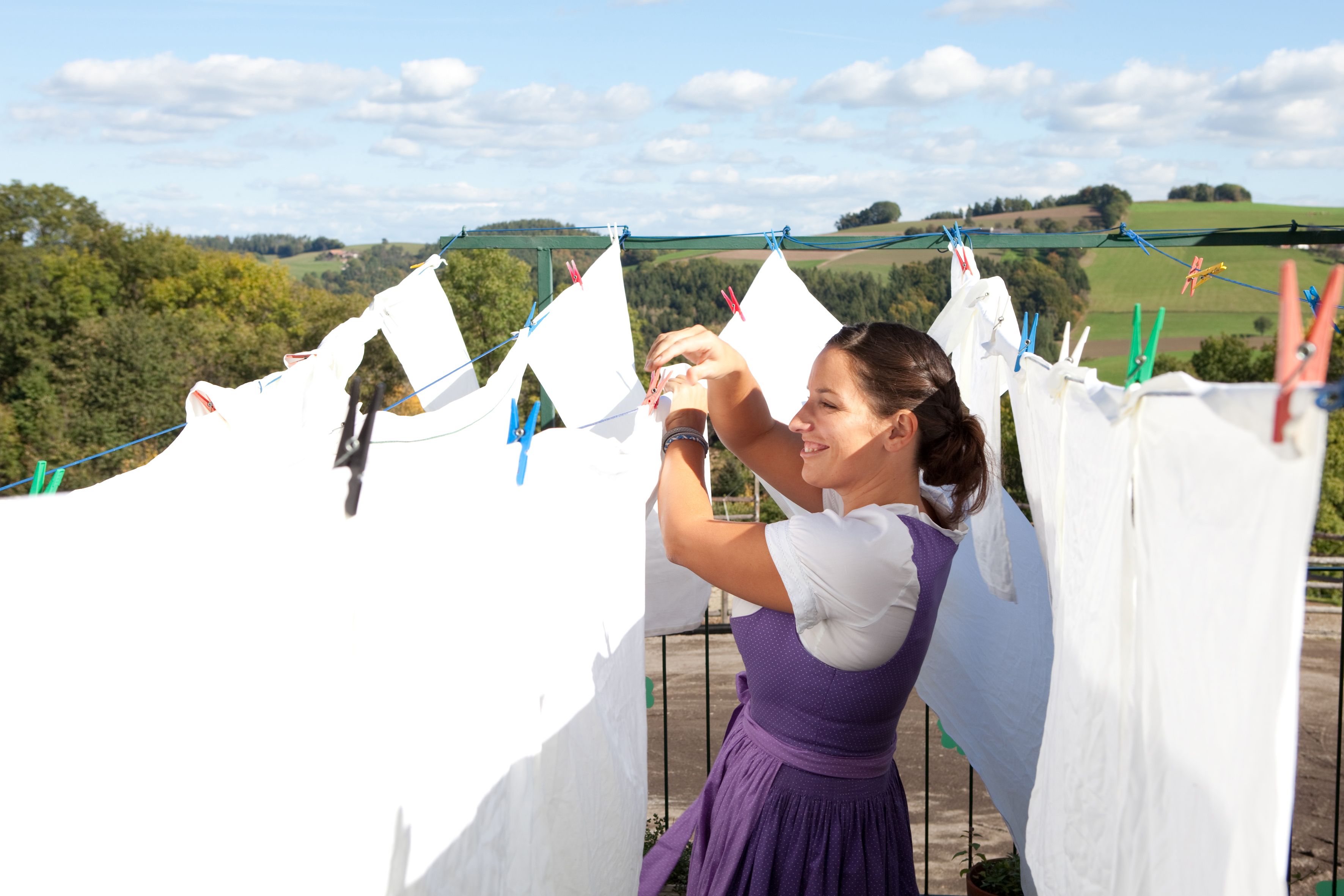 Woman hangs laundry outdoors, with rural landscape in the background.