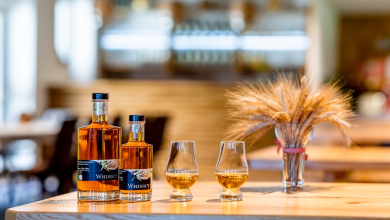 Two bottles of whisky and two glasses of whisky stand on a wooden table, with a bunch of cereals next to them.