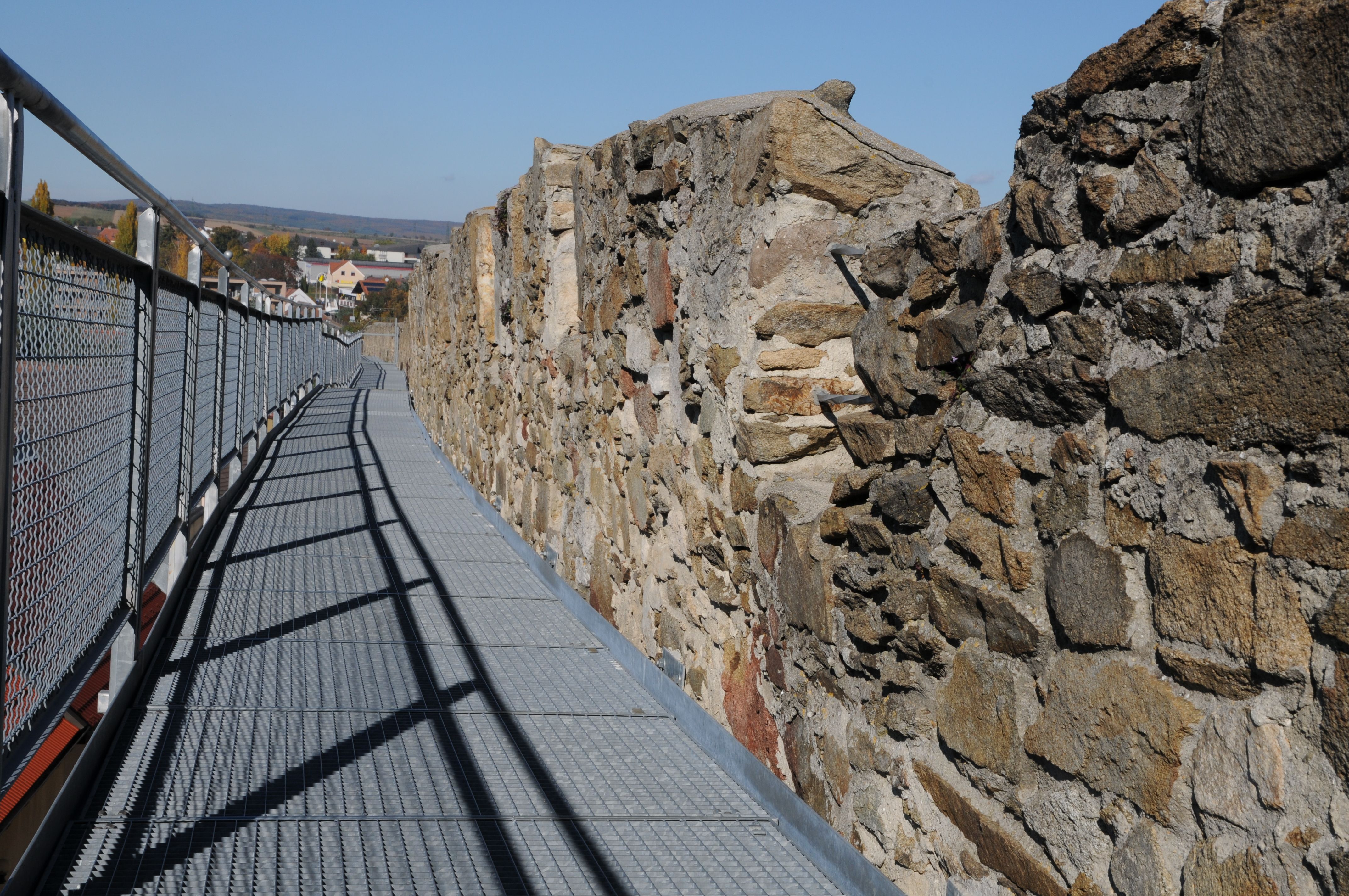A metal walkway along an old stone wall with a view of a town in the background.