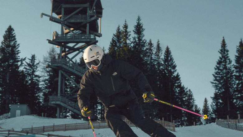 Skier in black clothing skiing on a groomed slope on the Semmering Hirschenkogel, with a lookout tower and trees in the background.