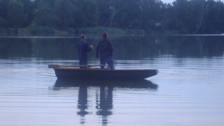 Two people fishing on a boat on a calm lake surrounded by trees.