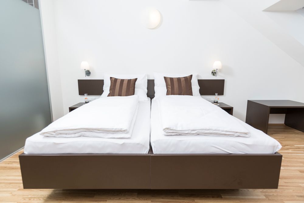 Modern guest room with two single beds, white comforters and brown pillows, wooden floor and bedside tables with lamps.