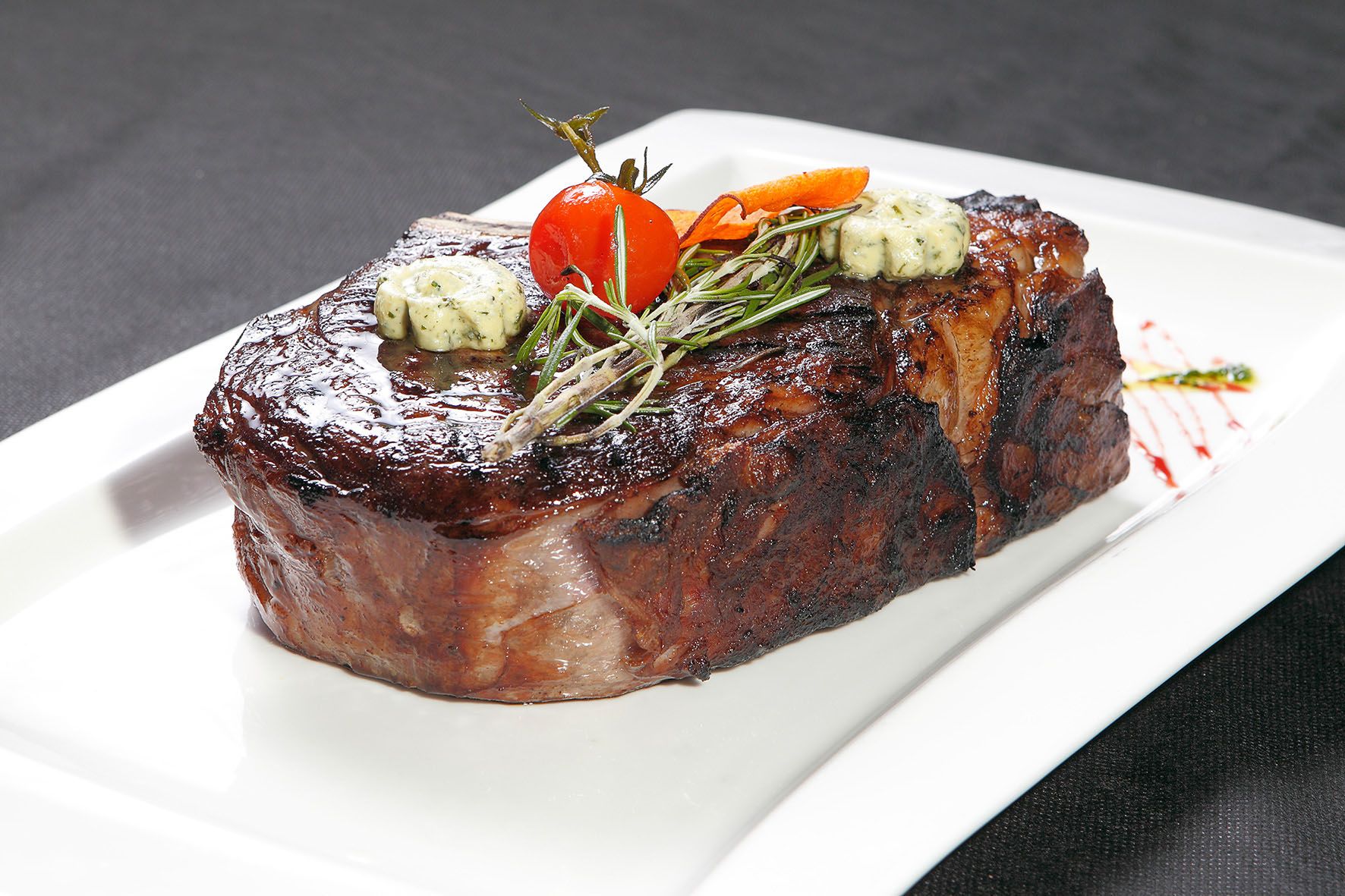 A juicy steak with herb butter, rosemary and a cherry tomato on a white plate.