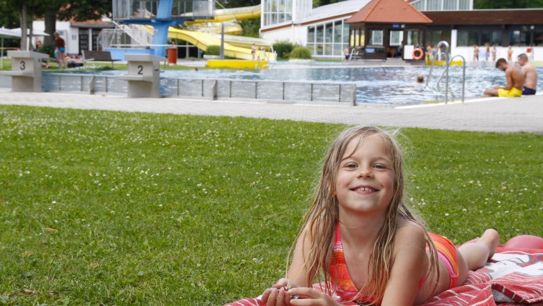 A girl lies on a red blanket in the grass in front of a swimming pool with slides and diving platform.