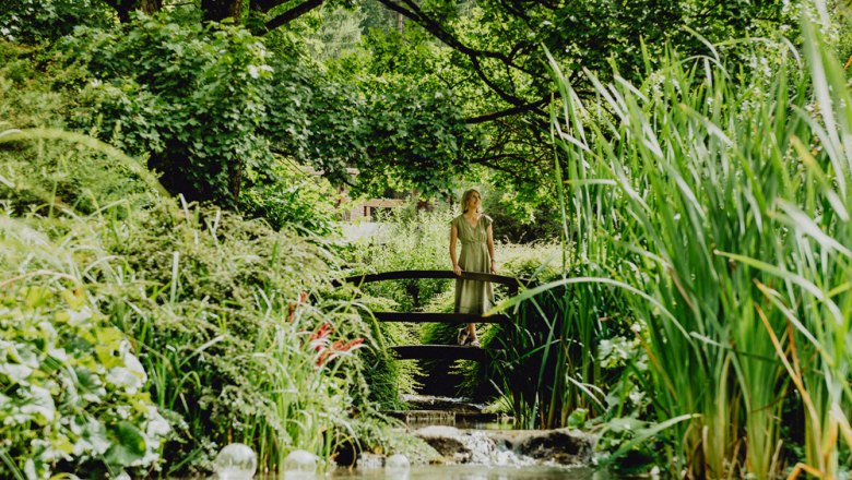 Woman standing on a wooden bridge in a lush, green garden with a stream.