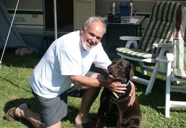 Animal lover, © L.G A man kneels next to a brown dog on a meadow in front of a caravan.