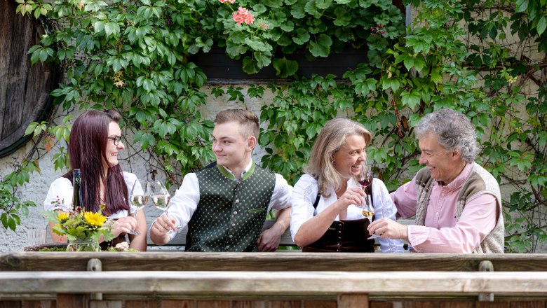 Four people in traditional dress clink glasses of wine outside in front of a wall covered in plants.