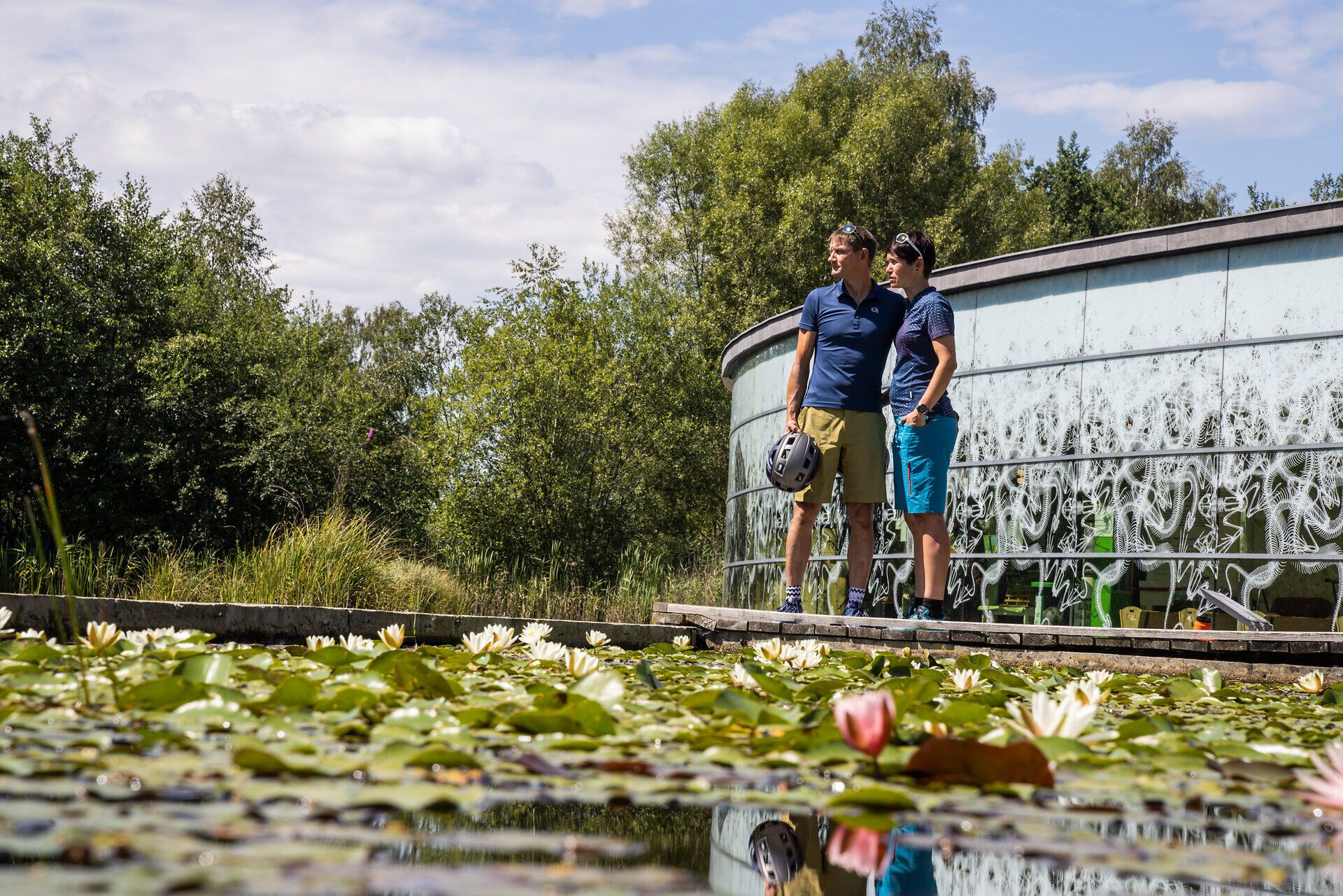 Two cyclists stand in the middle of a picturesque water landscape, enjoying the peace and beauty of nature. Surrounded by lush greenery and blooming water lilies, the sky is reflected in the clear water, while the gentle breeze creates a refreshing atmosphere.