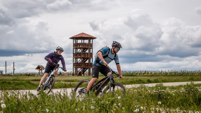 Two cyclists ride along a path in front of a wooden tower in a rural landscape.
