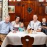 Family sitting at a table in front of a rustic wooden wall. There are 3 wine glasses and decorations on the table.