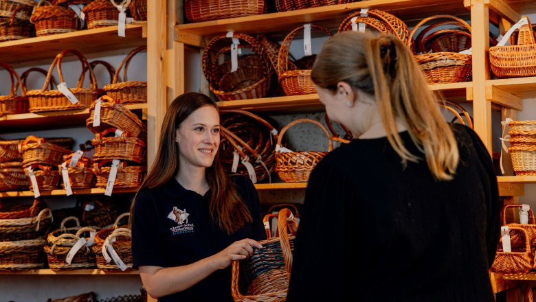 Two women in a store with woven baskets, one shows the other a basket.
