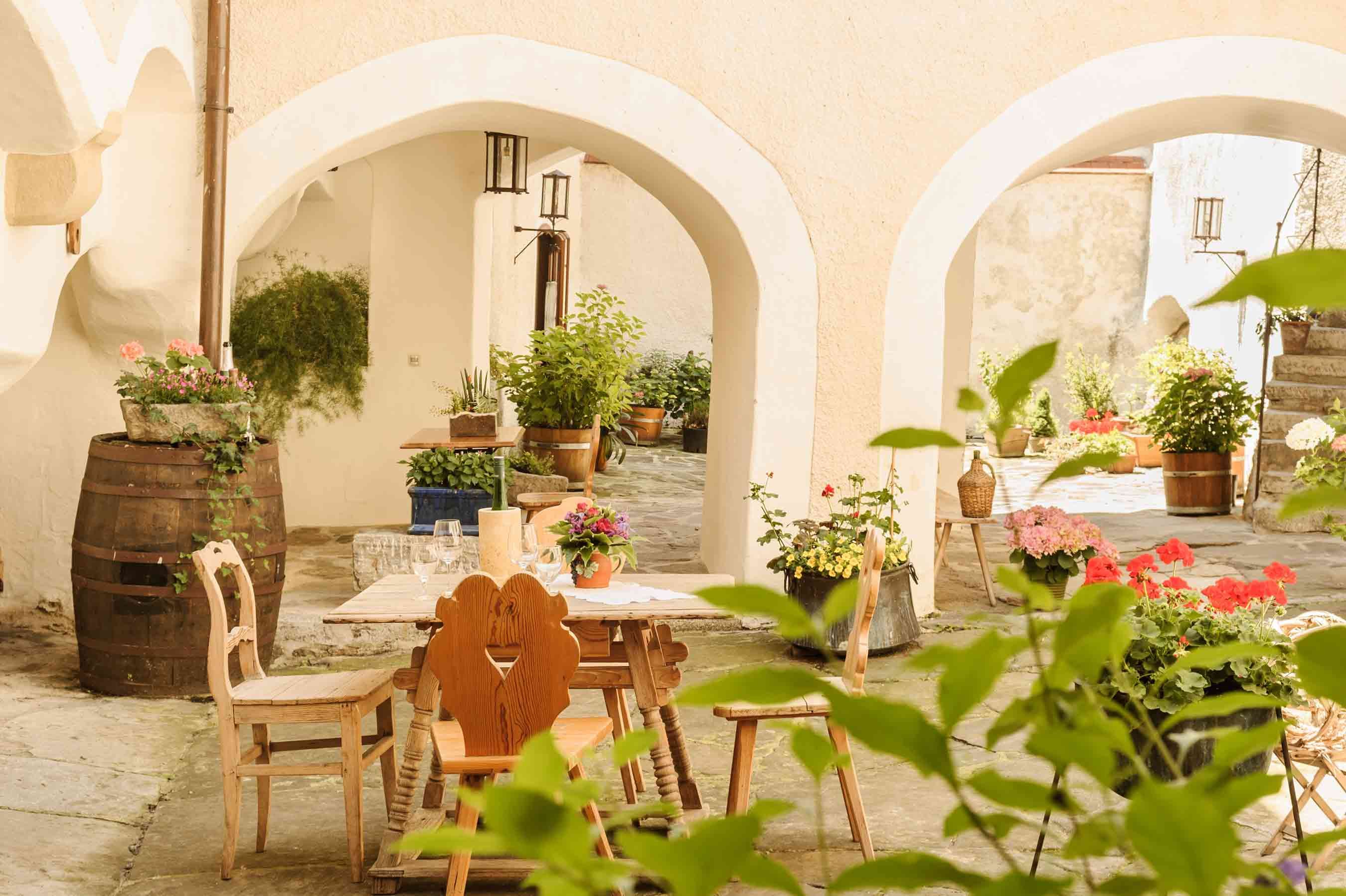 A cozy courtyard with a wooden table and chairs, surrounded by plants and flowers in pots.
