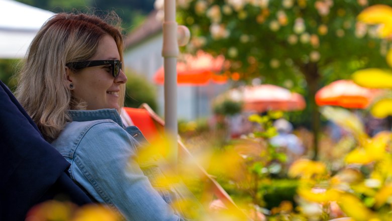 A woman lies on a deckchair under a parasol in the rose garden and smiles.
