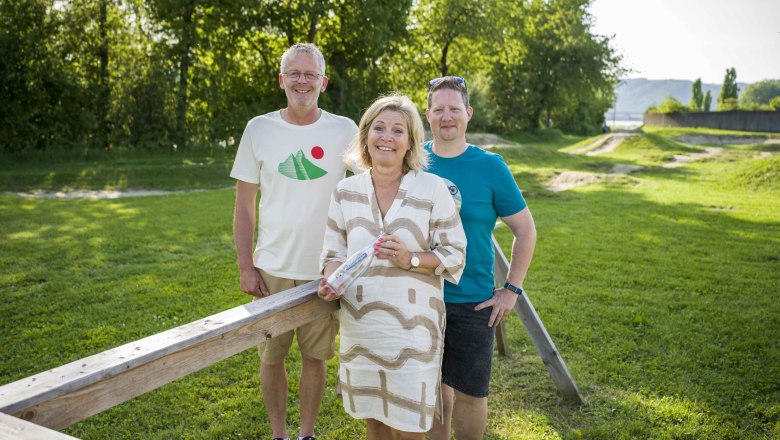 Three people are standing on a meadow in front of a wooden railing, surrounded by trees.