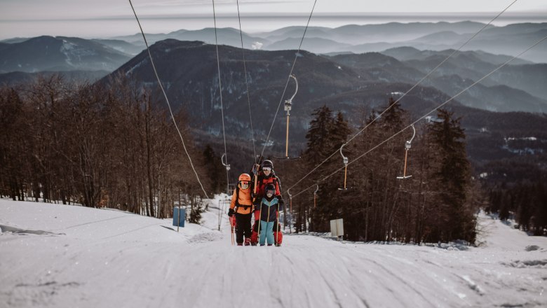 Three skiers on a drag lift in the Unterberg ski area, surrounded by snow-covered mountains and trees.