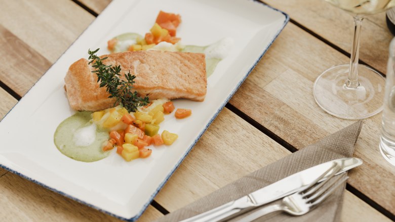 A plate with roasted salmon on vegetables and green sauce, decorated with a sprig of herbs, on a wooden table with cutlery and a wine glass.