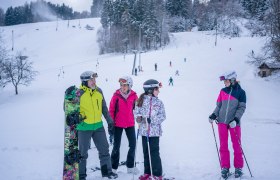 Group of skiers and snowboarders on a snowy slope with forest in the background.