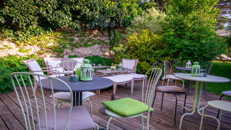 Garden area with tables and chairs on a wooden terrace, surrounded by green plants.