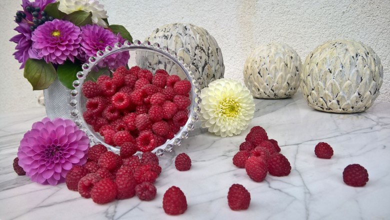 Raspberries in a bowl with flowers and decorative balls on a marble table.